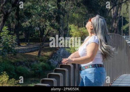 Ancienne touriste latino-américaine en profil debout sur un pont avec des arbres verts dans un fond flou, en profitant d'une promenade dans un ap public Banque D'Images