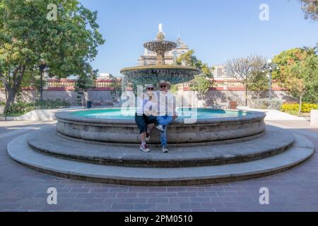 Paysage urbain de quelques touristes assis au bord d'une fontaine contre le ciel bleu, arbres verts et Parroquia de San Pedro Apostol en arrière-plan, plaza ne Banque D'Images