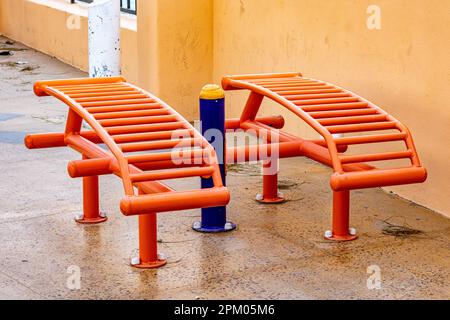 Deux banquettes tubulaires orange ouvertes s'assoient dans une salle de sport extérieure sur une promenade contre un mur jaune en arrière-plan, des feuilles sèches sur un sol humide, une journée nuageux à Baja Banque D'Images