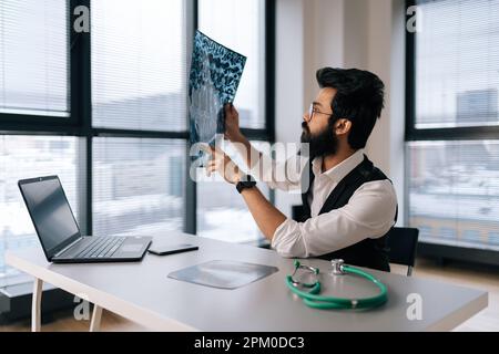 Portrait d'un médecin indien barbu orienté dans des lunettes pensant au diagnostic regardant les images IRM scanner par fenêtre. Banque D'Images