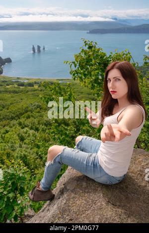 Femme bossy en T-shirt et jeans bleus assis sur la falaise sur la côte rocheuse dans les montagnes contre la vue panoramique sur la forêt verte et l'océan le jour d'été Banque D'Images