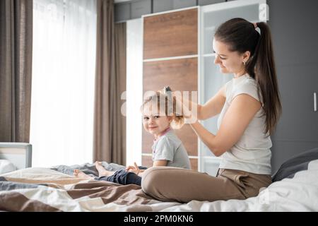 La jeune mère peigne les cheveux de sa petite fille tout en étant assise au lit le matin Banque D'Images