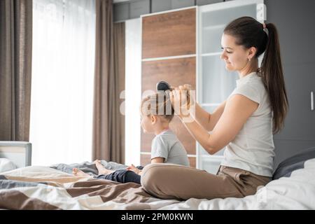 La jeune mère peigne les cheveux de sa petite fille tout en étant assise au lit le matin Banque D'Images