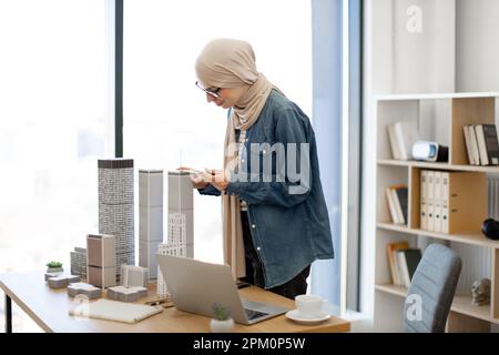 Bonne dame musulmane portant le hijab beige en utilisant la boussole de dessin tout en travaillant avec des modèles architecturaux au bureau. Professionnel de la conception efficace comparant les mesures avec les données numériques sur ordinateur. Banque D'Images