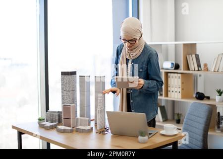 Jolie femme arabe en hijab beige remplaçant les modèles de bâtiment sur le bureau selon les informations sur l'appareil portable en main. Architecte efficace comparant les données du plan de projet à l'aide d'une tablette. Banque D'Images