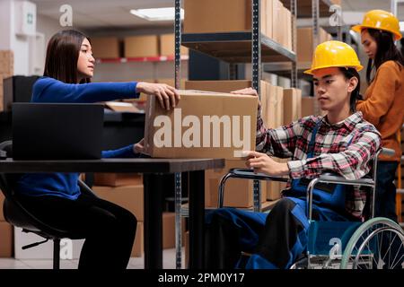 Opérateur d'entrepôt asiatique et préparateur de commande en fauteuil roulant, gérant la livraison des colis à la réception. Deux collègues du service postal traitant la boîte de carton de fret dans l'entrepôt Banque D'Images