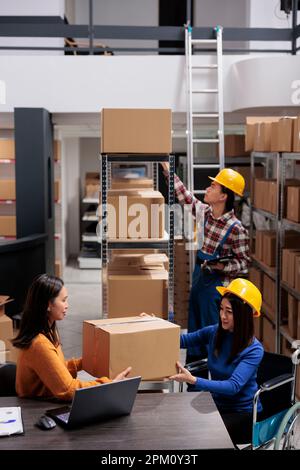 Le préparateur de commande d'entrepôt donne le colis du responsable prêt à être expédié. Une femme asiatique travaille en fauteuil roulant et un livreur s'occupe de la distribution des colis au bureau de la salle d'entreposage Banque D'Images