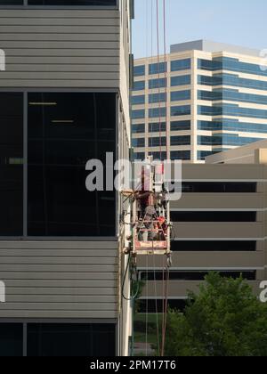 Vue à hauteur des yeux de deux lave-glaces mâles en uniforme qui nettoient le verre d'un immeuble de bureaux en se tenant sur un échafaudage suspendu. Banque D'Images