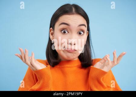 Gros plan d'une femme japonaise surprise, hausse et se répand les mains sur le côté, semble confus, se dresse sur fond bleu Banque D'Images