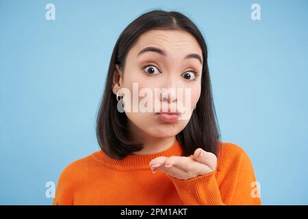 Gros plan de la belle femme asiatique envoie un baiser d'air à l'appareil photo, se dresse sur fond bleu Banque D'Images