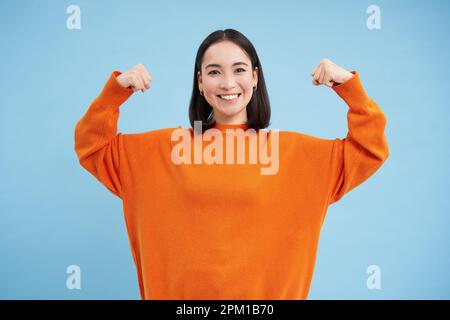 Portrait d'une femme coréenne mignonne en flexion de son biceps, montre des bras forts, des muscles et des sourires, concept de personnes en bonne santé et en forme, fond bleu Banque D'Images