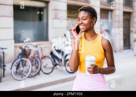 Belle femme africaine debout dans la rue et souriante en parlant par téléphone. Elle porte une tenue décontractée d'été et une coupe de cheveux tendance Banque D'Images