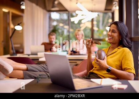Femme d'affaires mature avec des pieds sur le bureau travaillant sur l'ordinateur portable dans le bureau parlant dans le micro du téléphone mobile Banque D'Images