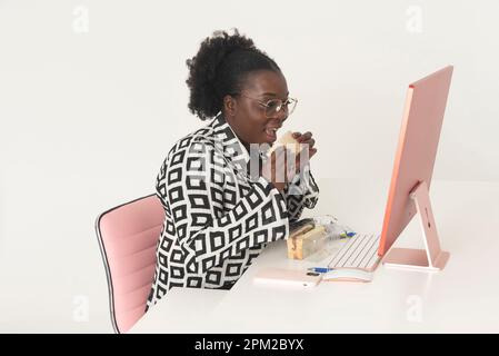Angleterre, Royaume-Uni. 2023. Travailleur de bureau noir ayant un sandwich pour le déjeuner tout en travaillant sur un ordinateur. Banque D'Images