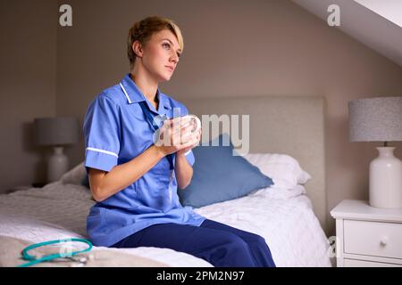 Femme réfléchie portant un uniforme d'infirmières ou de médecins avec boisson chaude assise sur le lit à la maison Banque D'Images