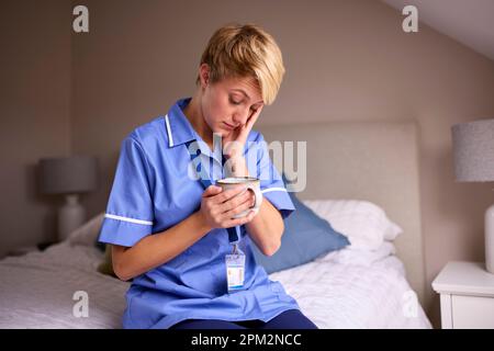Femme fatiguée portant des infirmières ou des médecins uniforme avec boisson chaude assis sur le lit à la maison Banque D'Images
