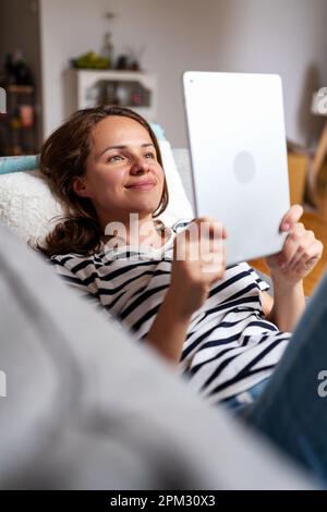 Une femme brune se trouve sur un canapé et regarde des vidéos de médias sociaux sur Internet à l'aide de sa tablette numérique. Banque D'Images