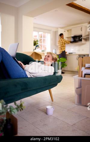 Couple Déballage des boîtes après avoir déplacé dans la nouvelle maison ensemble comme la femme se trouve sur le canapé en utilisant l'ordinateur portable Banque D'Images