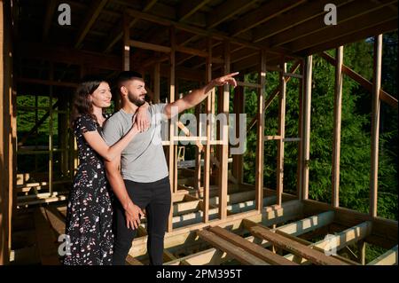 Homme et femme inspectant leur future maison de cadre en bois nichée dans les montagnes près de la forêt. Jeune couple sur le chantier en début de matinée. Concept de construction écologique contemporaine. Banque D'Images