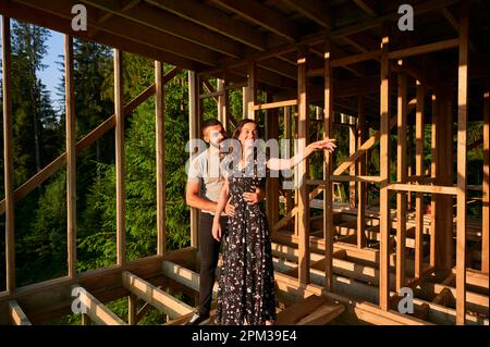 Homme et femme inspectant leur future maison de cadre en bois nichée près de la forêt. Jeune couple sur le chantier en début de matinée. Concept de construction écologique contemporaine. Banque D'Images