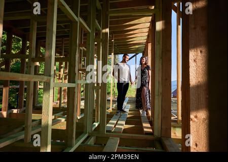 Homme et femme inspectant leur future maison de cadre en bois nichée près de la forêt. Jeune couple sur le chantier en début de matinée. Concept de construction écologique contemporaine. Banque D'Images