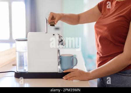 Vue latérale d'une femme insérant une capsule de café dans une machine à espresso dans la cuisine Banque D'Images