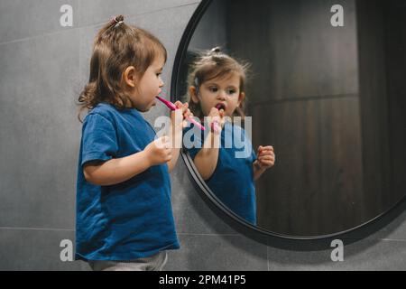 Bébé fille se brossant les dents avec la brosse à dents comme la routine quotidienne des enfants et la procédure d'auto-soin, regardant dans le miroir. Hygiène dentaire des enfants. Soins de santé pour enfants. Banque D'Images