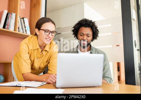 Deux collègues heureux et divers hommes afro-américains et caucasiens regardant l'écran d'ordinateur portable et assis dans le hall du bureau Banque D'Images