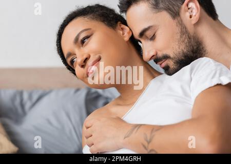 une femme afro-américaine souriante et rêveuse qui regarde près d'un homme barbu qui l'embrasse dans sa chambre à la maison, photo Banque D'Images