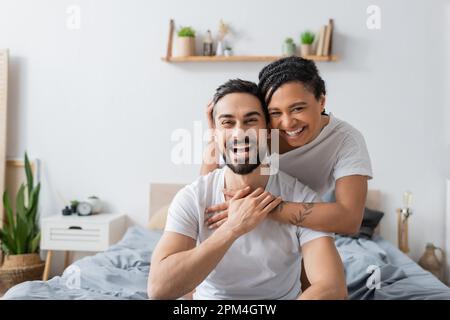 un couple multiethnique débordé en t-shirts blancs riant à la caméra dans la chambre à la maison, image de stock Banque D'Images