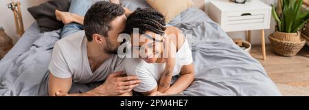 homme joyeux qui dit secret à la jeune femme afro-américaine tout en se reposant sur le lit à la maison, bannière, image de stock Banque D'Images