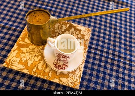 Une casserole en métal (cezve) de café turc mousseux se trouve ici à côté d'une petite tasse sur une tuile décorée au Boutique Hotel Hanna Koumi à Kormakitis (Korucam), République turque de Chypre du Nord (RTNC). Pour un arôme complet, le café est bouilli trois fois, une épaisse couche de mousse étant considérée comme essentielle. Banque D'Images
