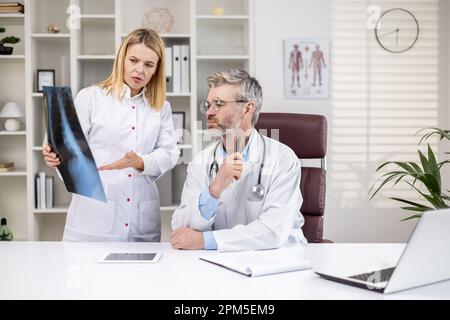 Médecins sérieux et matures pensant et discutant de la radiographie du patient, homme et femme en couches blanches médicales travaillant à l'intérieur du bureau de la clinique, consultation de travail. Banque D'Images