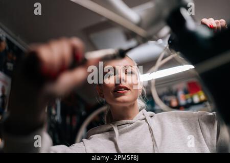 Vue rapprochée à angle bas de la jolie femme blonde mécanique de vélo de réparation et de fixation vélo de montagne travaillant dans l'atelier de réparation de vélo avec obscurité Banque D'Images