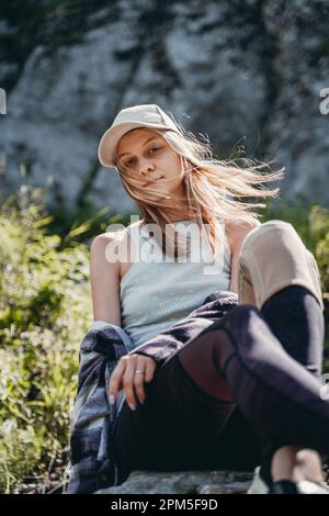 une fille dans un haut gris et une casquette blanche est assise sur une pierre Banque D'Images