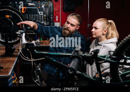 Vue avant du réparateur barbu en vélo communiquant avec la cliente blonde, parlant du problème de vélo détecté pendant le diagnostic dans Banque D'Images