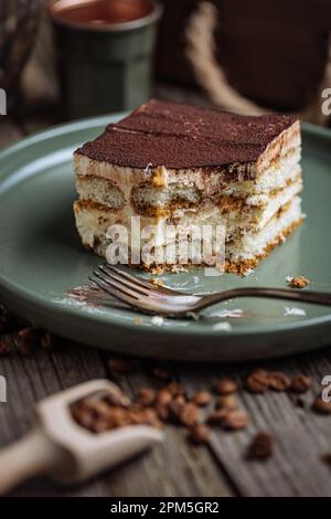 Un morceau de gâteau Tiramisu, dessert italien traditionnel, servi dans une ambiance rustique et de style moody. Fond en bois et plaque verte. Banque D'Images