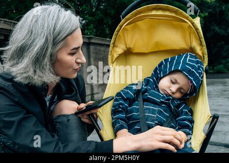 La femme regarde l'enfant endormi assis dans la poussette avec le téléphone dans les mains Banque D'Images