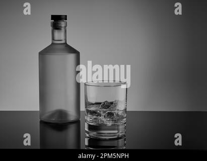 Image en noir et blanc d'une bouteille de spiritueux avec un verre à whisky avec de la glace et de l'alcool shooté sur une surface réfléchissante. Banque D'Images