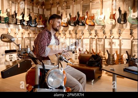 Joyeux souriant jouant de la guitare acoustique assis à moto à la boutique de musique Banque D'Images