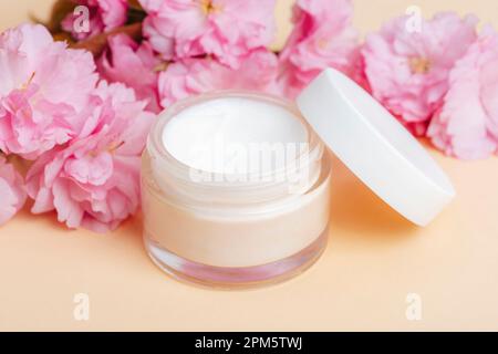 Crème visage dans un pot et fleur de cerisier rose sur fond beige. Soin de la peau, concept de traitement de beauté. Gros plan. Banque D'Images