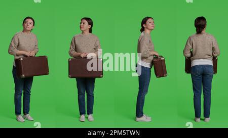 Modèle féminin tenant une valise à bagages sur l'appareil photo, se préparant à partir en voyage avec une mallette et des bagages. Femme caucasienne portant un sac de voyage vintage debout sur fond de plein corps de greensscreen. Banque D'Images