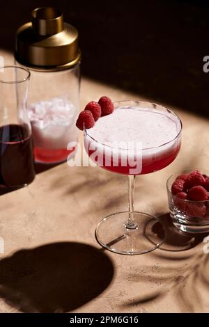 Cocktail de club de trèfle dans un grand verre coupé garni de trois framboises à côté d'un shaker en lumière dure. Banque D'Images