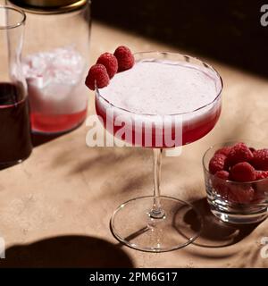 Cocktail de club de trèfle dans un grand verre coupé garni de trois framboises à côté d'un shaker en lumière dure. Banque D'Images