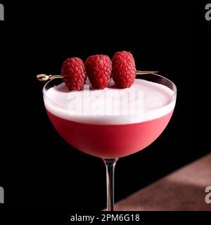 Cocktail de club de trèfle dans un grand coupé en verre garni de trois framboises sur une sélection d'or. Banque D'Images