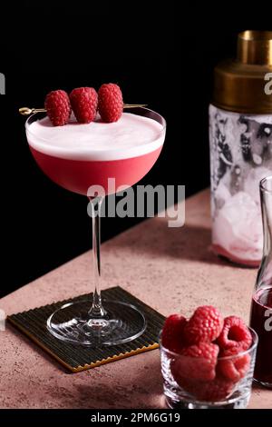 Cocktail de club de trèfle dans un grand coupé en verre garni de trois framboises sur une sélection d'or. Banque D'Images