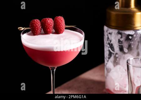 Cocktail de club de trèfle dans un grand coupé en verre garni de trois framboises sur une sélection d'or. Banque D'Images