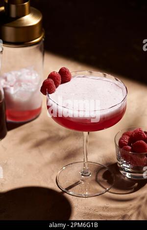 Cocktail de club de trèfle dans un grand verre coupé garni de trois framboises à côté d'un shaker en lumière dure. Banque D'Images