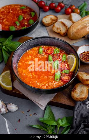 Soupe de riz aux tomates garnie de feuilles de basilic, tomates cerises et une tranche de citron dans un bol noir, tranches de pain, feuilles de basilic et un autre bol de sou Banque D'Images