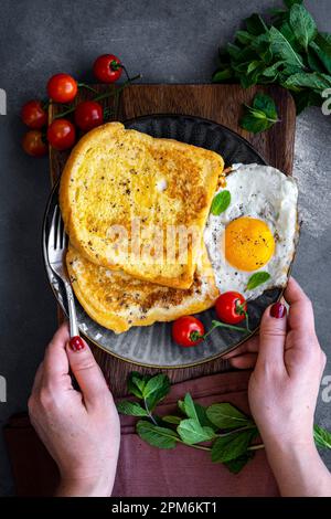 Mains tenant une fourchette et une assiette de petit-déjeuner chargée de tranches de pain grillé, d'œuf frit et de tomates cerises. Feuilles de menthe fraîche sur le côté. Banque D'Images